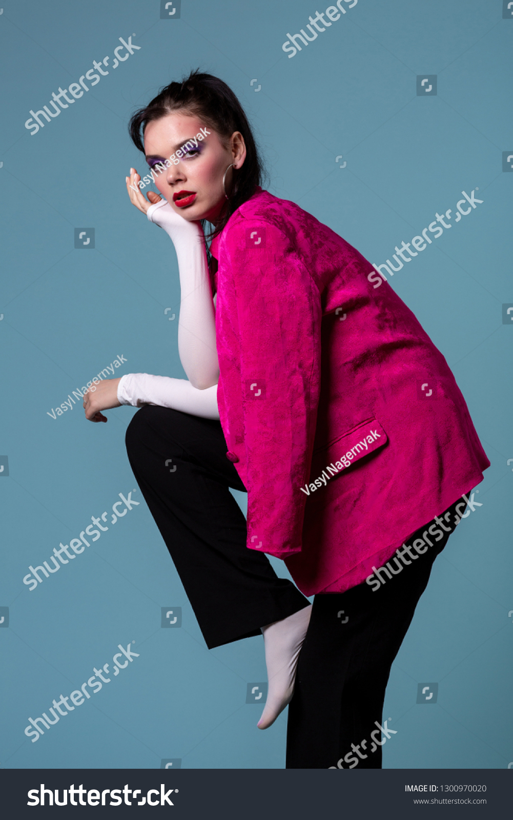 Amazing young brunett woman in trendy pink jacket standing on one leg, looking at the camera posing in the studio on blue background #1300970020