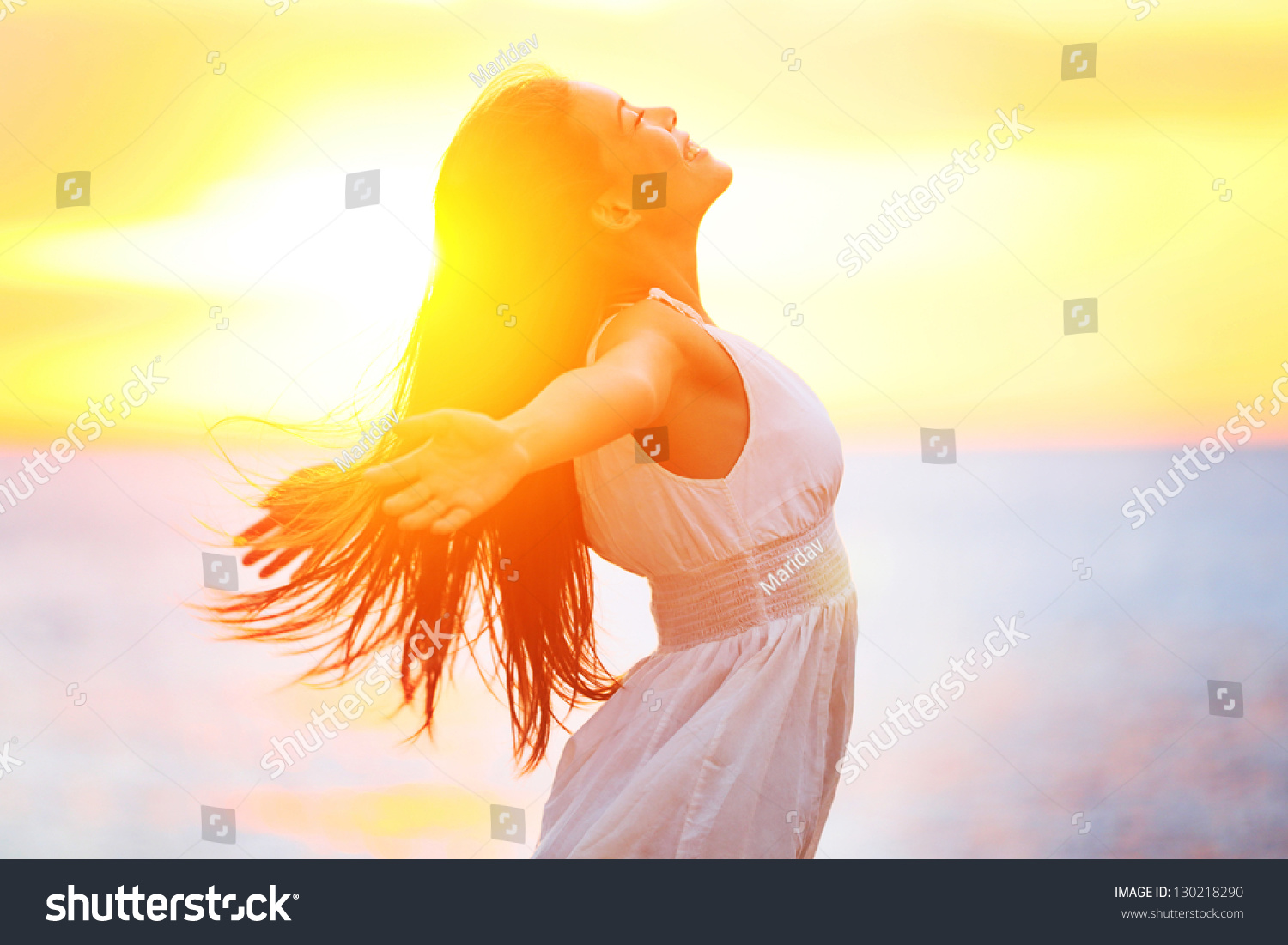 Enjoyment - free happy woman enjoying sunset. Beautiful woman in white dress embracing the golden sunshine glow of sunset with arms outspread and face raised in sky enjoying peace, serenity in nature #130218290