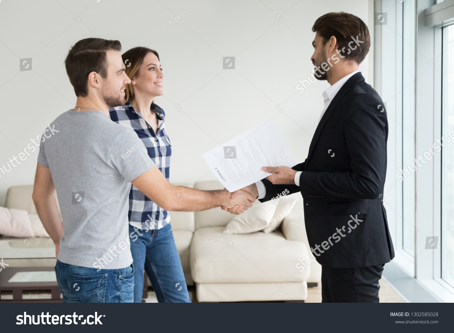 Realtor or landlord handshaking couple buyers tenants make real estate deal holding rental agreement or sale purchase contract, agent and clients shake hands welcoming renters in new home apartment #1302585028