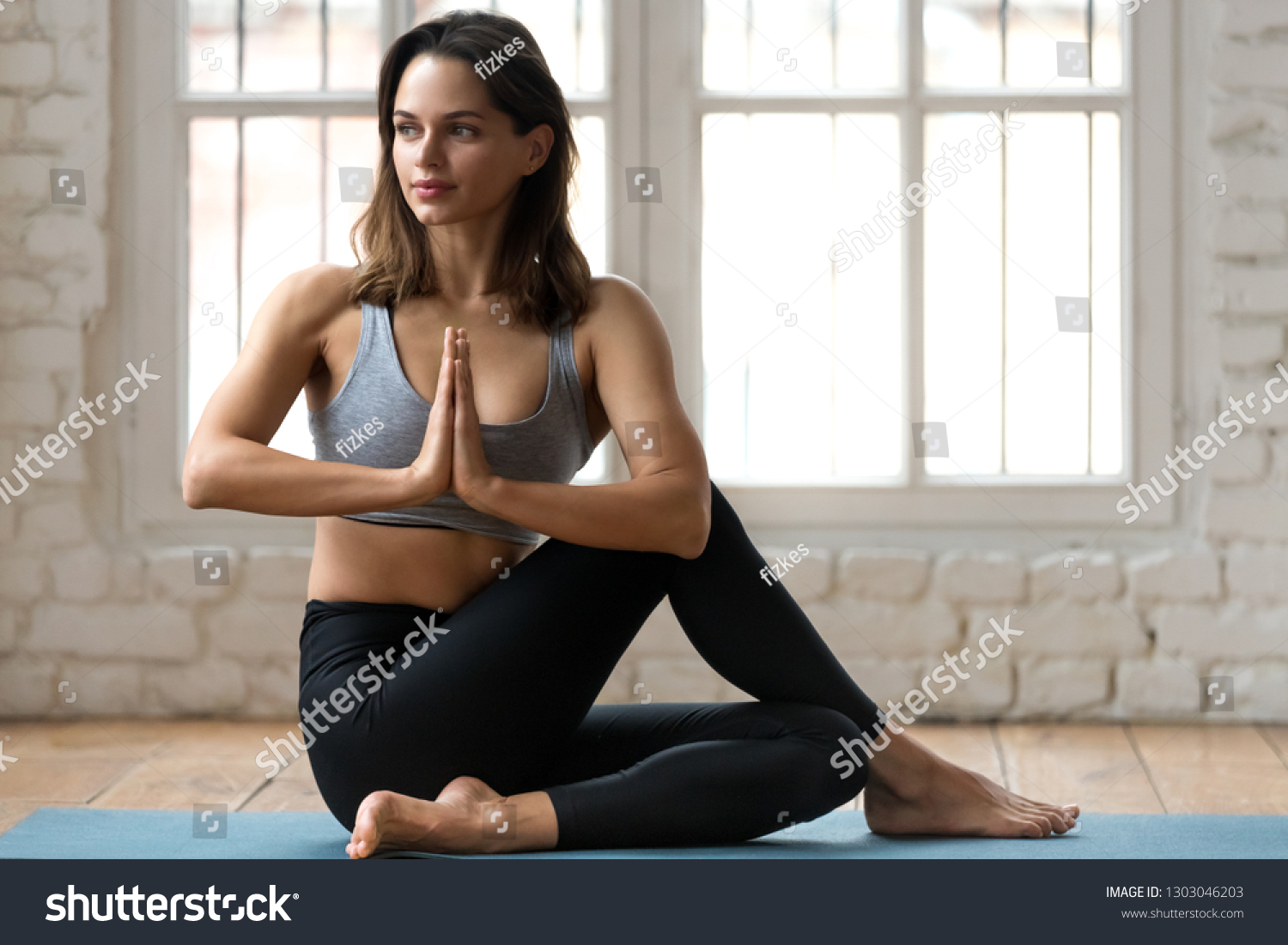 Young attractive woman practicing yoga, doing Half lord of the fishes exercise, Ardha Matsyendrasana pose with namaste , working out, wearing sportswear, pants and top, indoor full length, yoga studio #1303046203