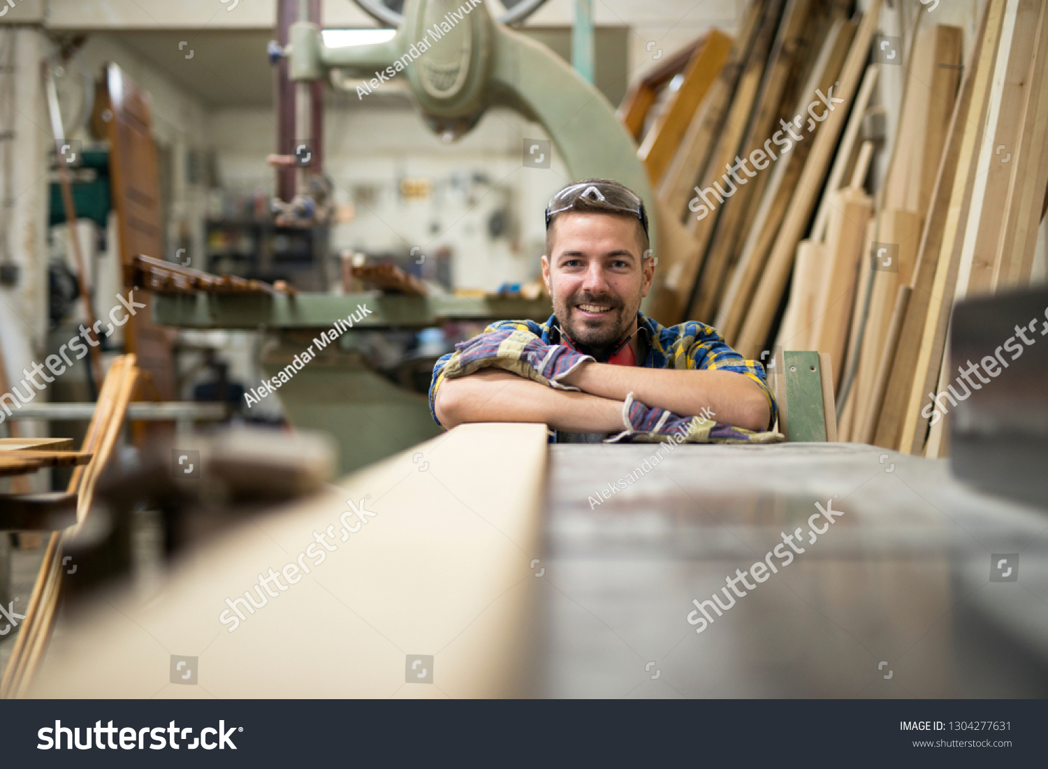 Portrait of professional woodworker standing next to a machine and wood material in his carpentry workshop. #1304277631
