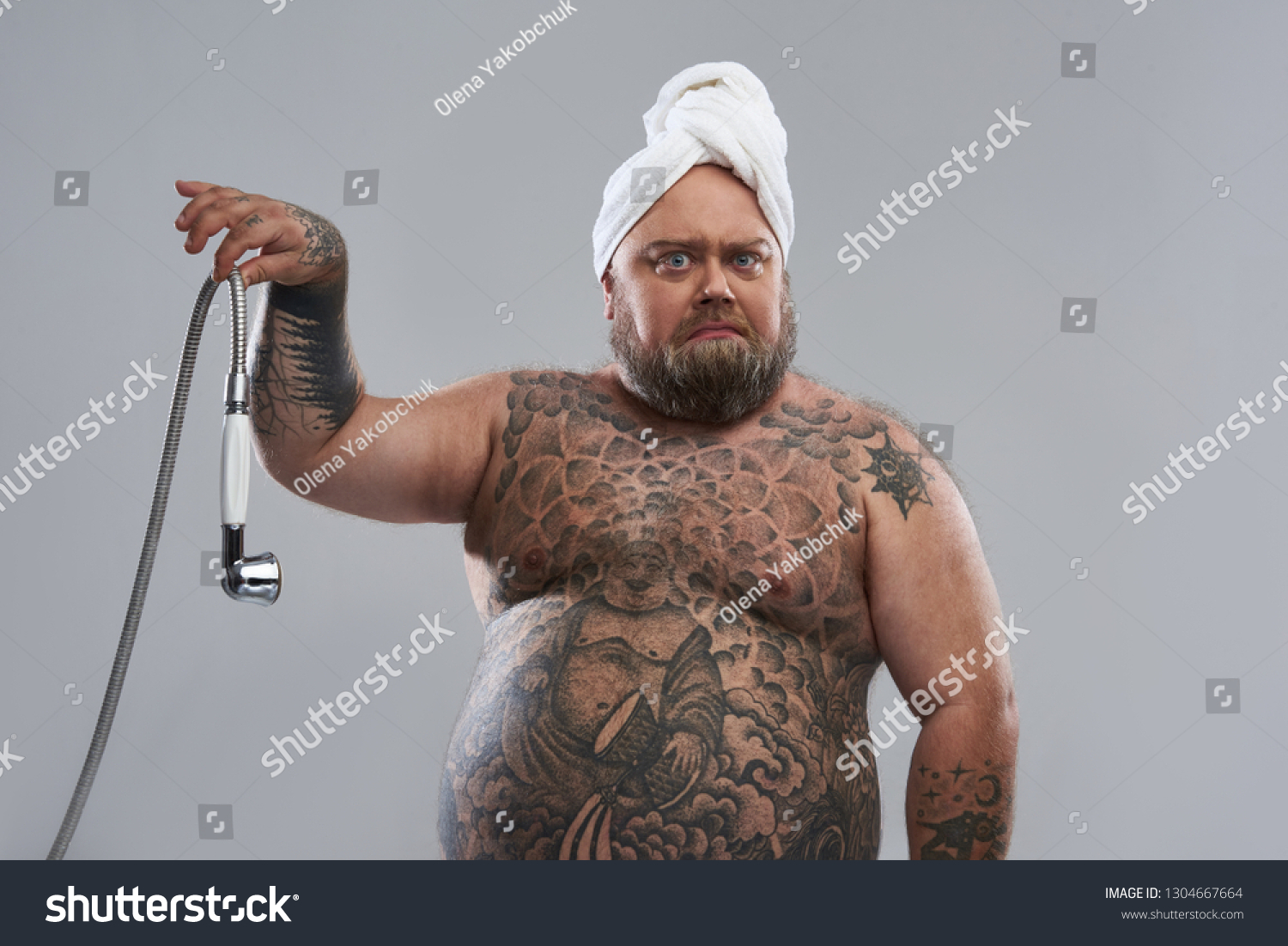 Tattooed fat man with towel on his head standing isolated on the grey background and frowning while holding shower hose with two fingers #1304667664