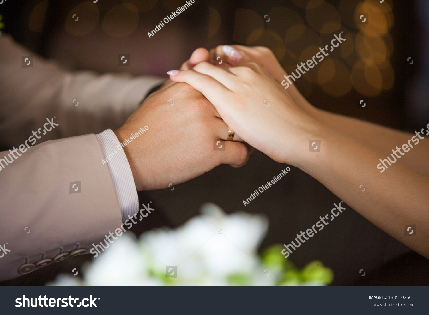 Close up side view of man and woman holding hands while sitting at wooden table in front of each other. Female and male hands of bride and groom on wedding day. Horizontal color photography. #1305102661