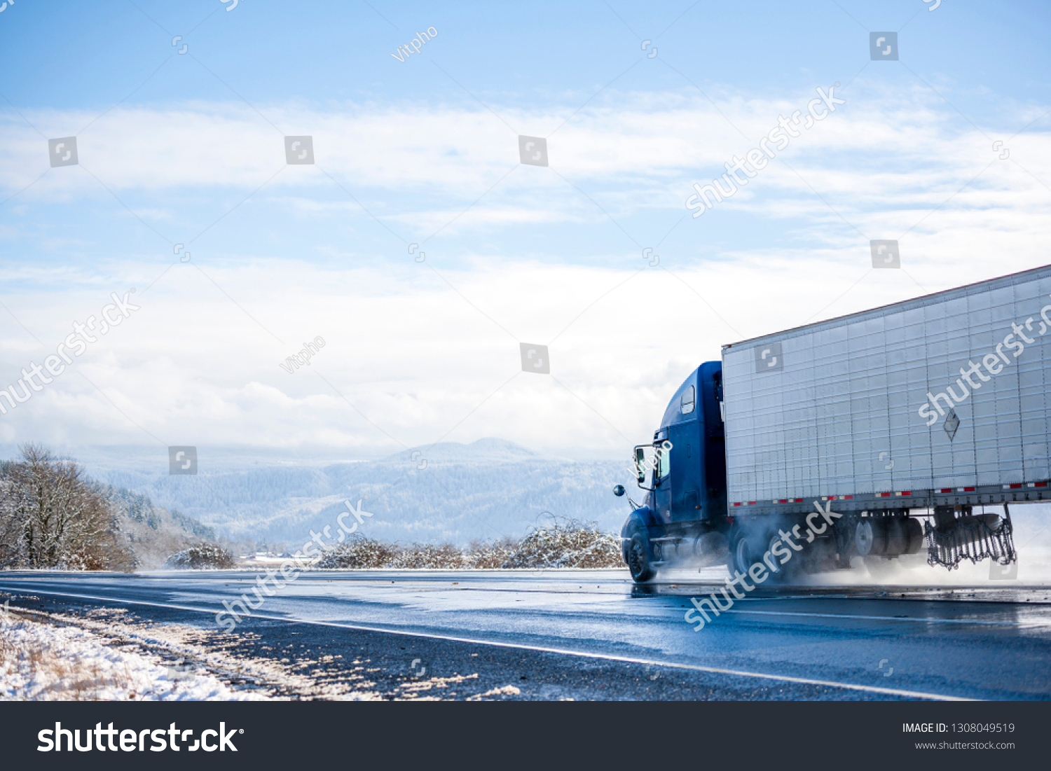 Big rig pro long haul blue semi truck tractor transporting commercial cargo in refrigerator semi trailer going on the wet glossy road with water from melting snow and winter snowy trees on the side #1308049519