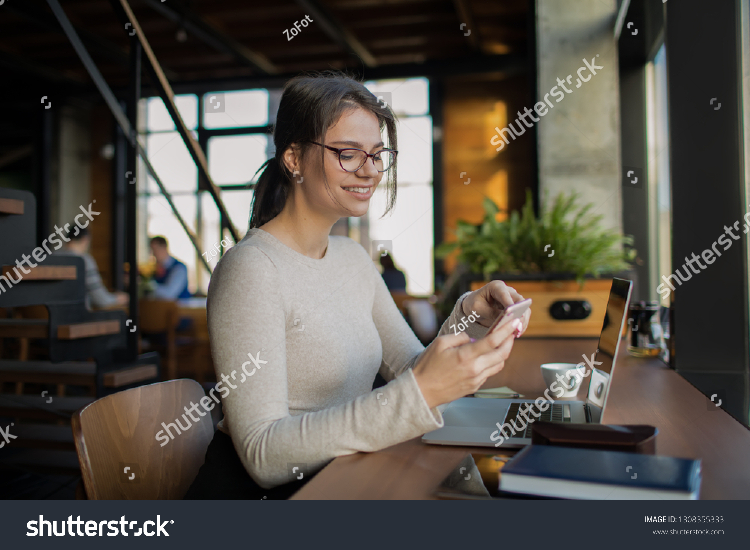 Happy smiling woman clever university student reading pleasant text message on smartphone during online learning on pc laptop computer, sitting at desktop in modern coffee shop. Online shopping store #1308355333