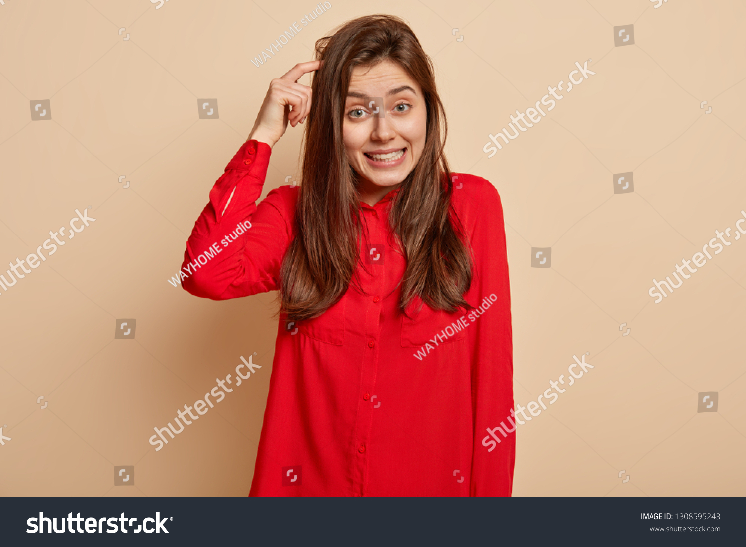 Puzzled unsure delighted young woman scratches head and looks directly at camera, makes decision, recieves good proposal, wears red shirt, poses over light beige background, uses imagination #1308595243