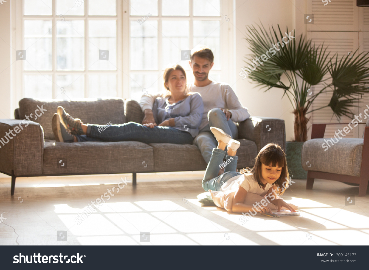 Happy parents relaxing on couch in comfort light living room while little kid child daughter playing on warm floor drawing with colored pencils, family having fun together, underfloor heating concept #1309145173