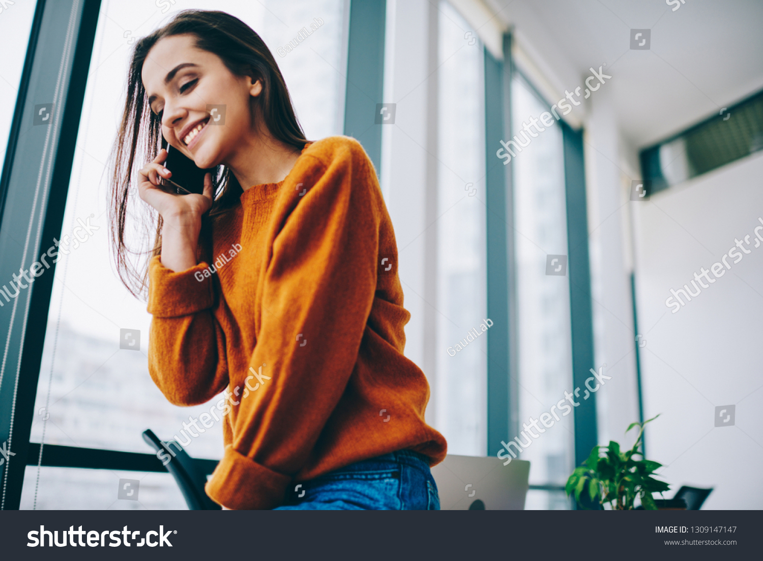Pretty caucasian employee using smartphone gadget for discussing information with friend connected to 4g internet indoors, happy cheerful hipster girl making mobile conversation via application #1309147147