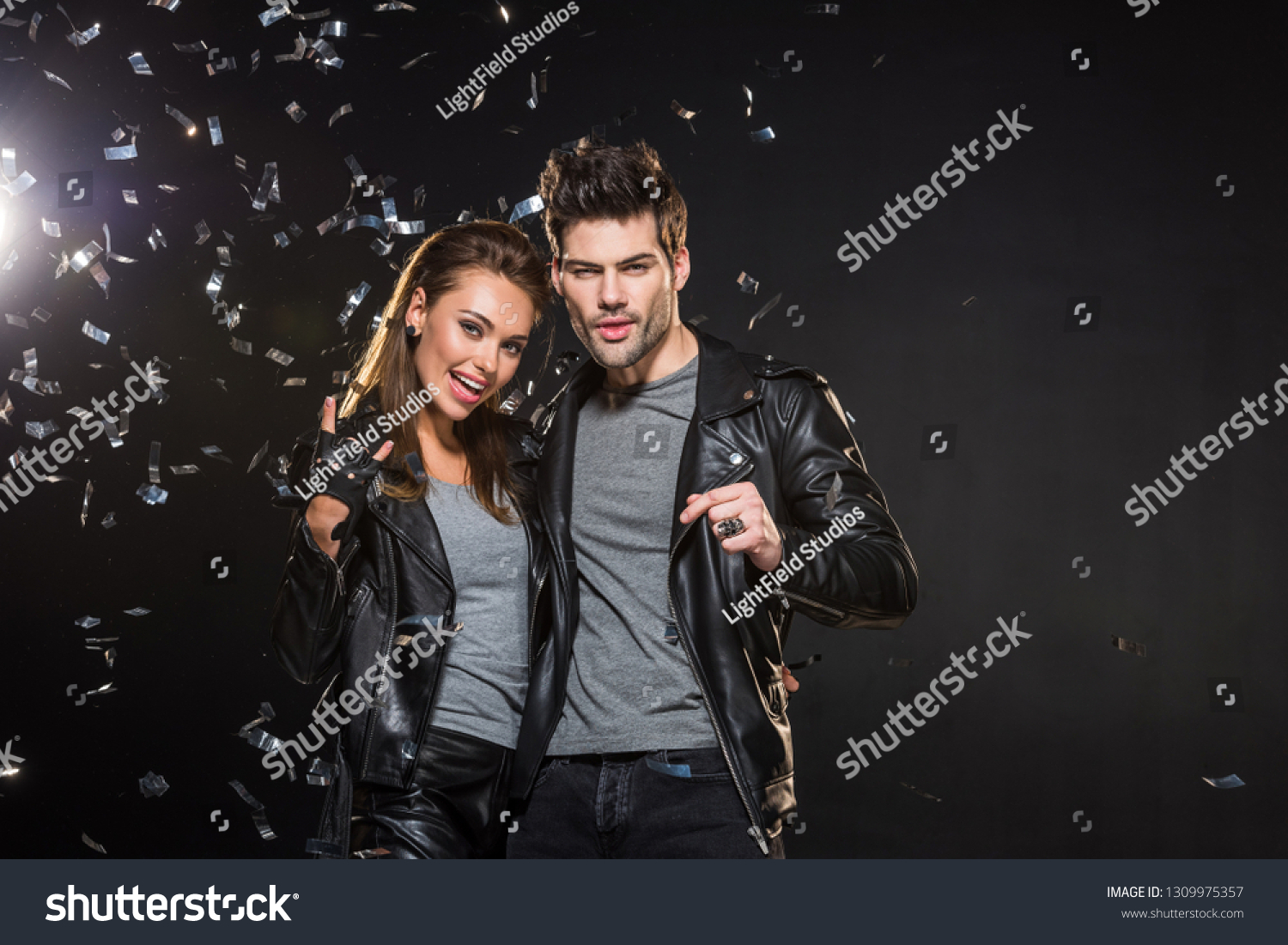 beautiful couple in leather jackets cheering and looking at camera with falling confetti on black background #1309975357