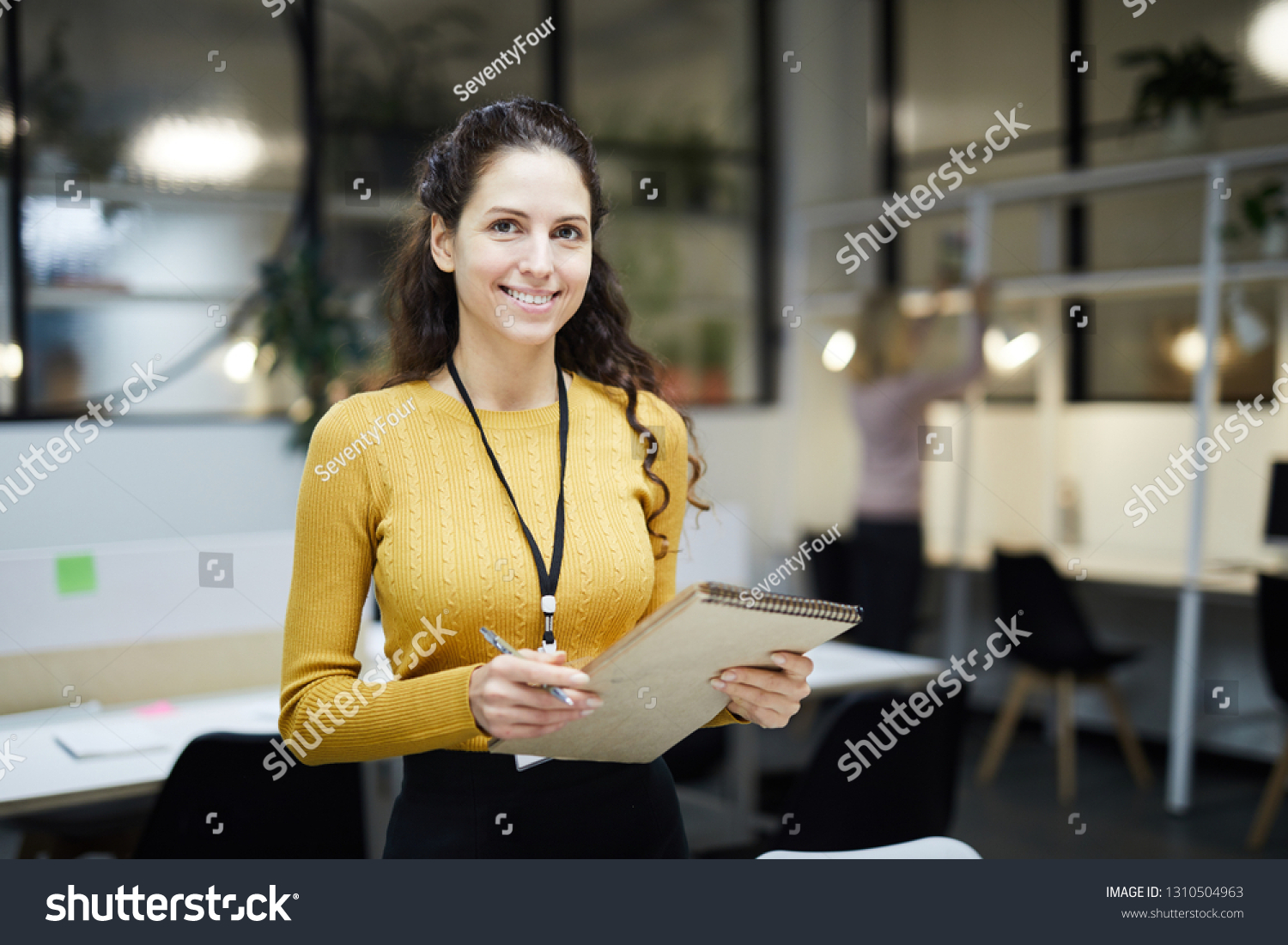 Smiling pretty young lady with badge on neck standing in modern open space office and holding sketchpad while looking at camera #1310504963