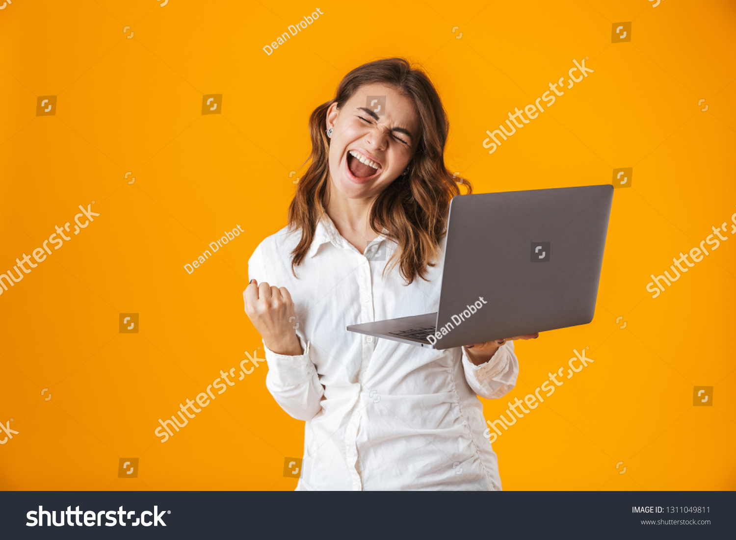 Portrait of a cheerful young woman wearing white shirt standing isolated over yellow background, holding laptop #1311049811