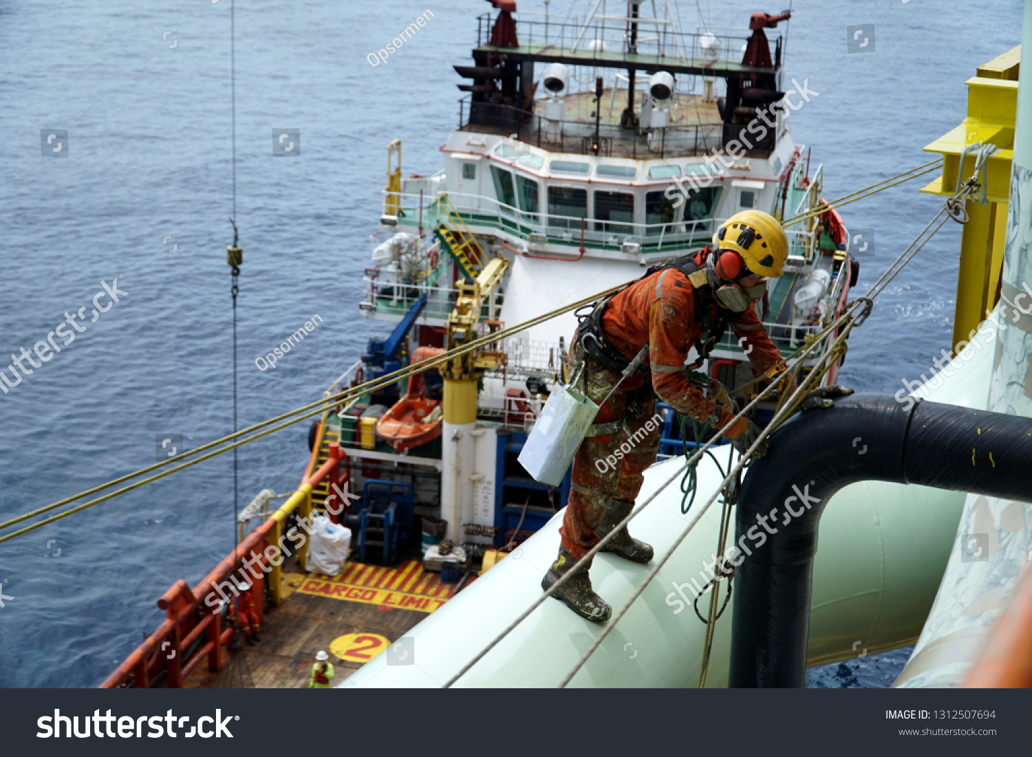 An abseiler wearing Personal Protective Equipment (PPE) standing on pipeline for painting activities with background marine vessel floating at the sea. #1312507694