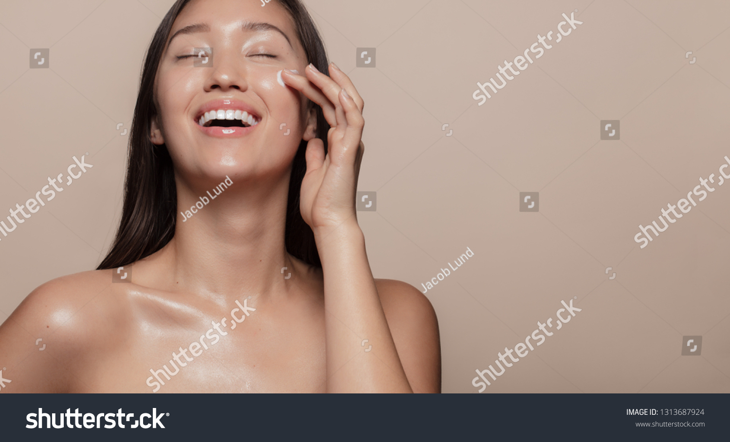 Beautiful girl with bare shoulders applying cream on her face and smiling against beige background. Smiling asian woman with glowing skin applying facial skincare cream with eyes closed.