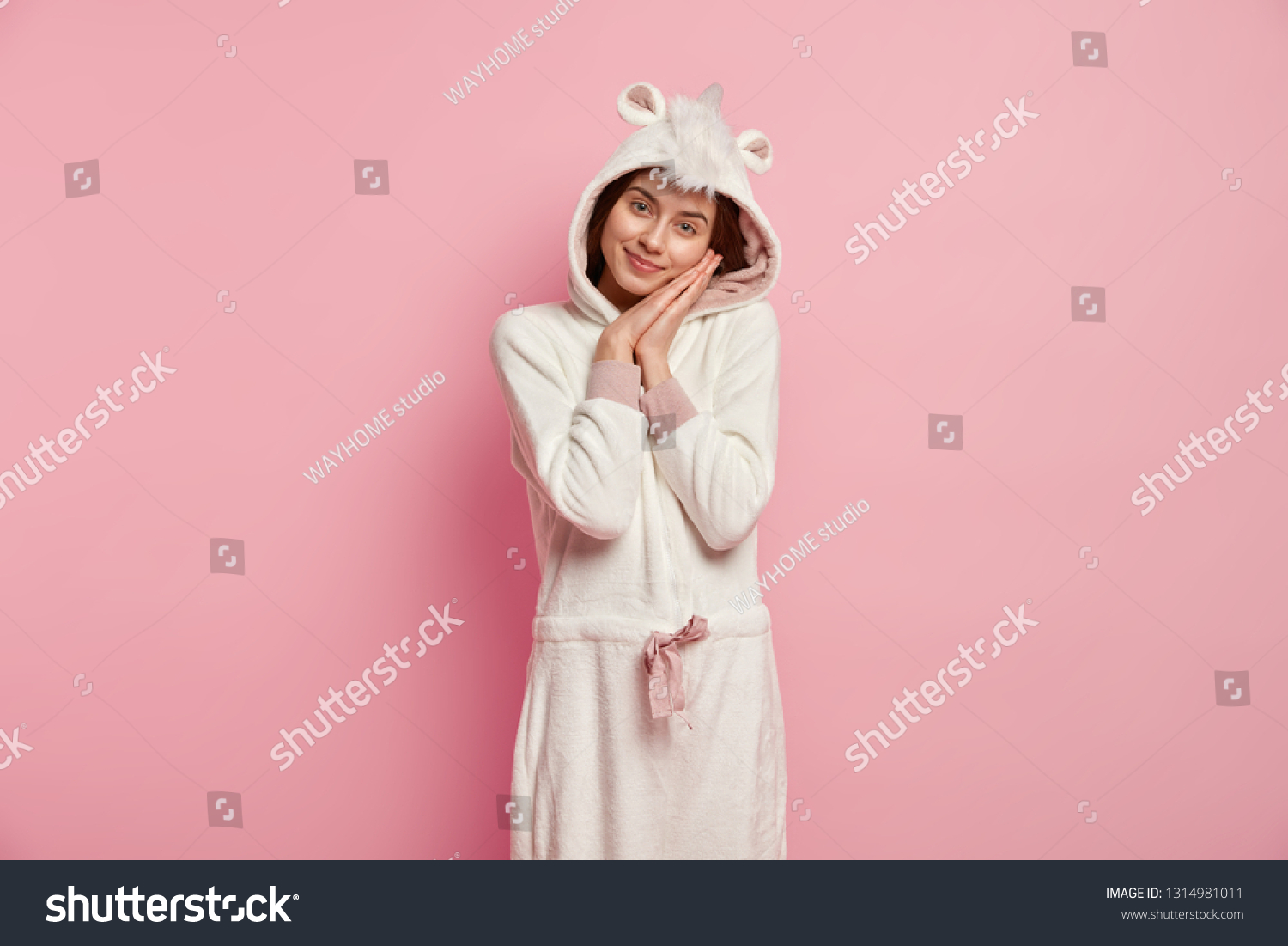 Restful girl leans at hands, enjoys good sleep, wears funny soft domestic outfit, looks at camera with tender look, poses over pink background, has nice rest in calm atmosphere. Bedding concept #1314981011