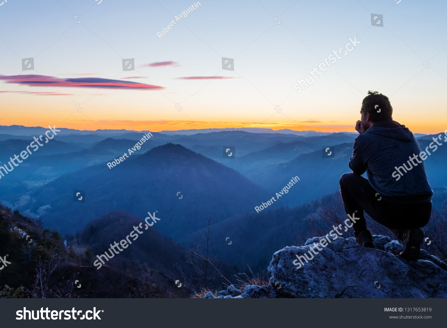 Male hiker crouching on top of the hill and enjoying scenic view of twilight landscape below. Hiking, achievement, expectation, optimism and self-reflection concepts. #1317653819