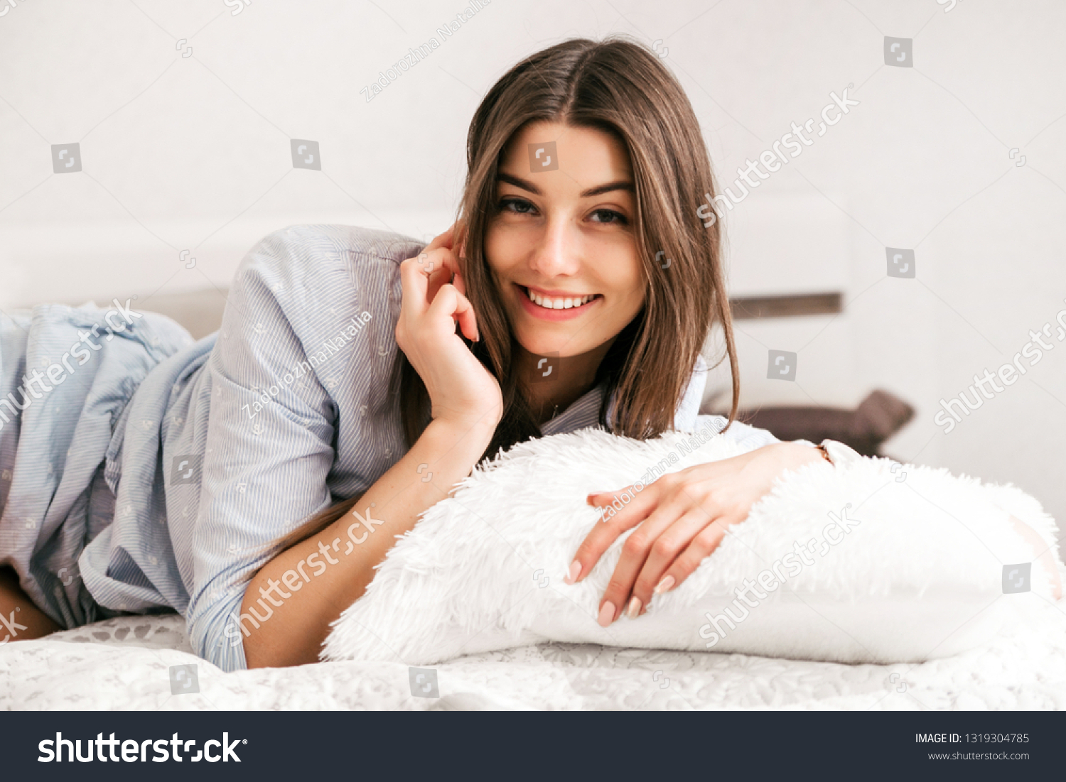 Young smiling woman lying in white bed and using a phone in her bedroom. Happy morning. #1319304785