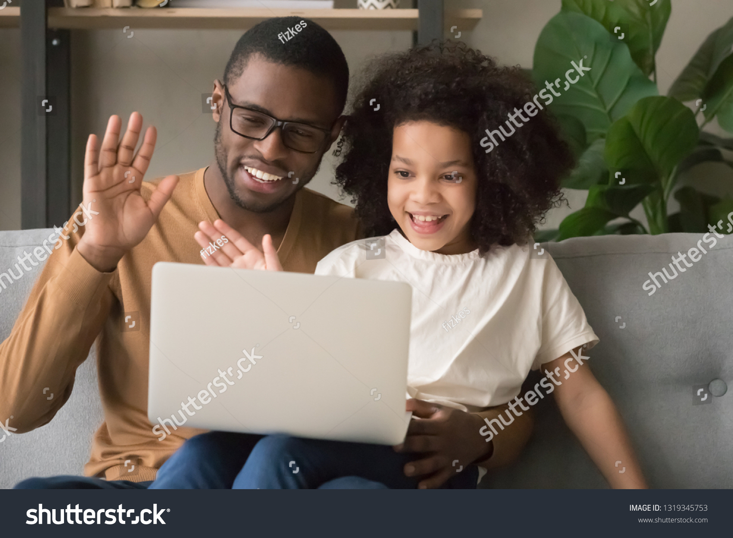 Happy family black dad with kid daughter waving hands making distance video call looking at laptop, african father and child girl looking talking to webcamera chatting online by computer webcam #1319345753