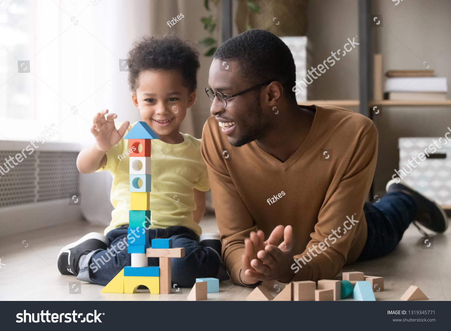 Happy cute little son playing game with black dad baby sitter building constructor tower from multicolored wooden blocks, african family father and toddler child boy having fun on warm floor at home #1319345771