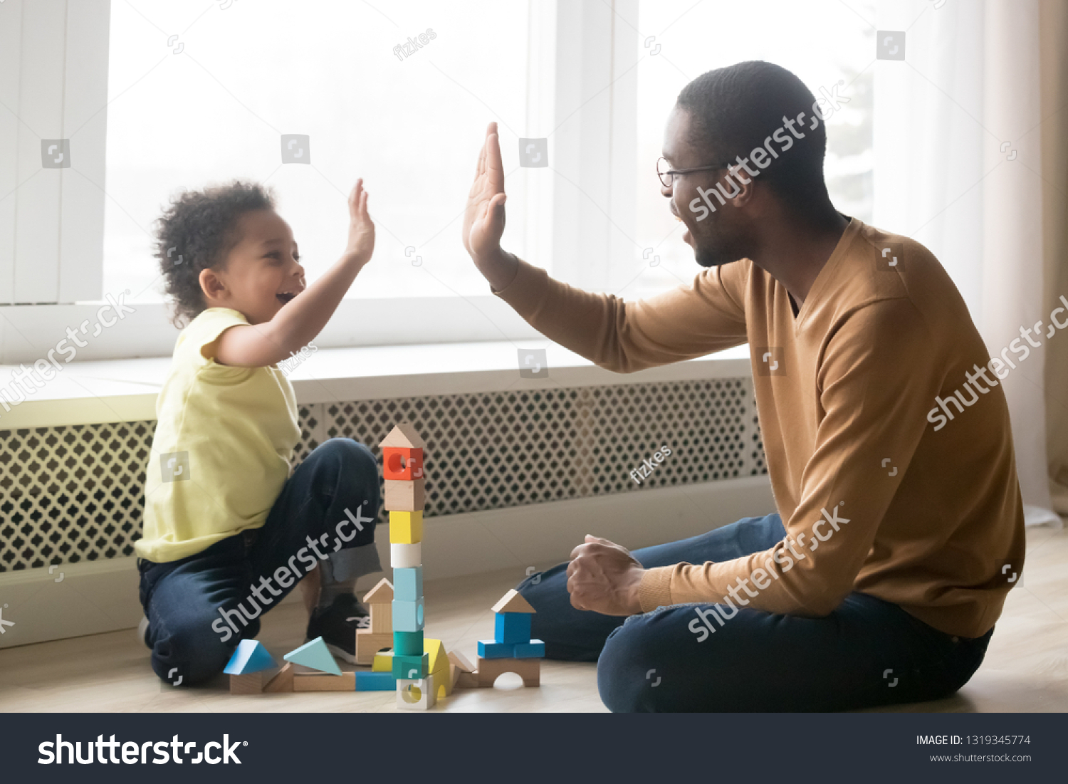 Happy black dad and cute toddler son giving high five playing with wooden blocks on floor at home, little kid boy enjoying game with african father, daddy child clapping hands having fun together #1319345774