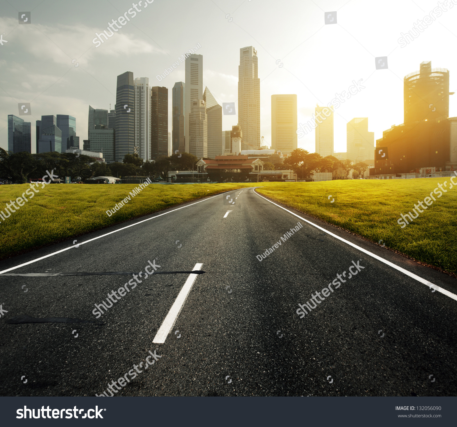 Asphalt road leading to a city with tall buildings through green meadow #132056090