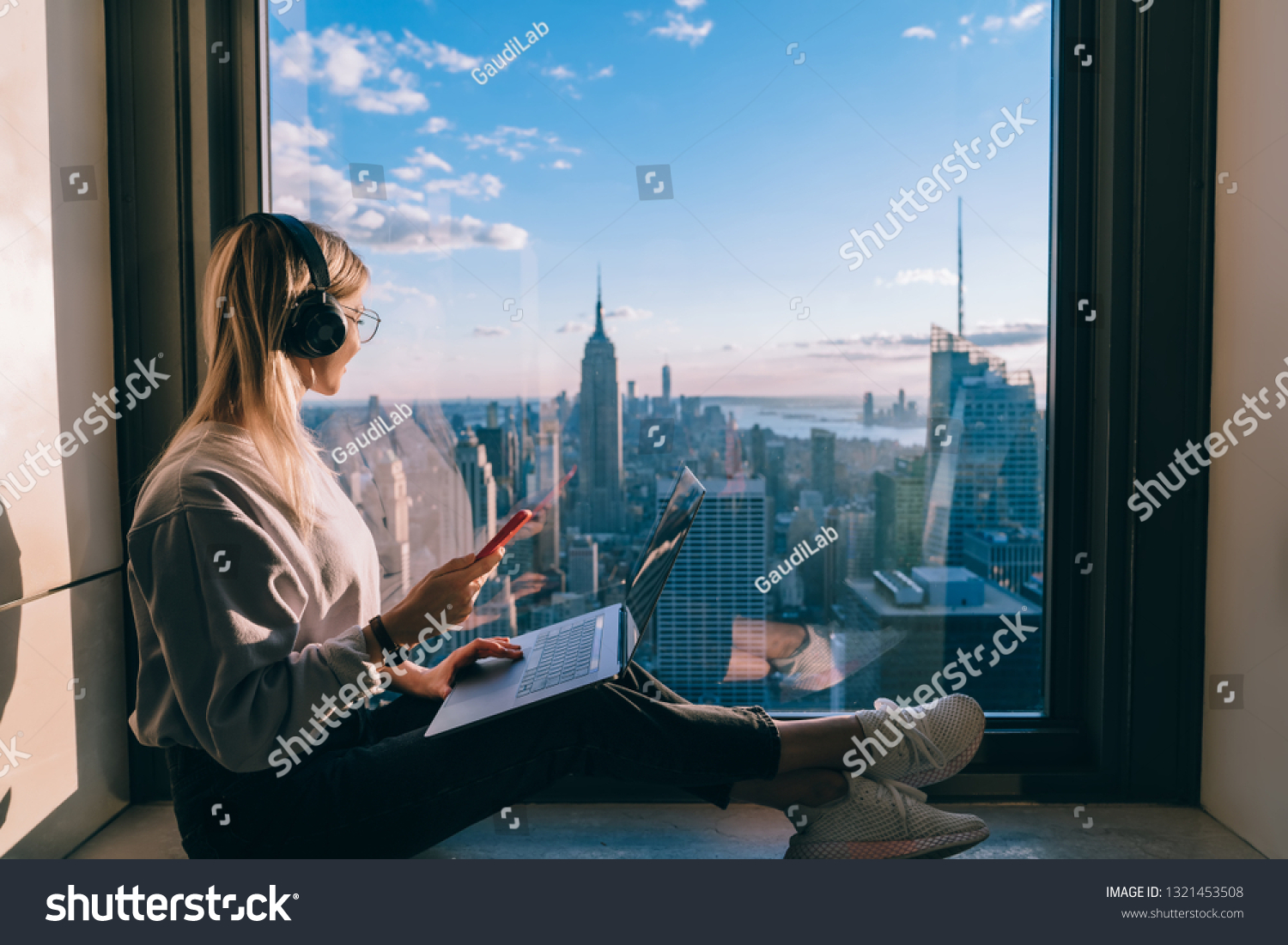 Blonde hair female blogger listening to music in headphones during freelance work on laptop computer. Hipster girl traveler enjoying panoramic view of New York downtown and studying with audio book #1321453508