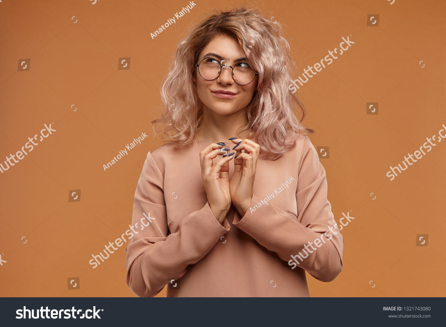 Sly hipster girl planning prank or evil trick, clasping hands and smiling mysteriously. Pensive cunning young woman in eyewear having tricky plan in mind, posing isolated at blank orange studio wall #1321743080
