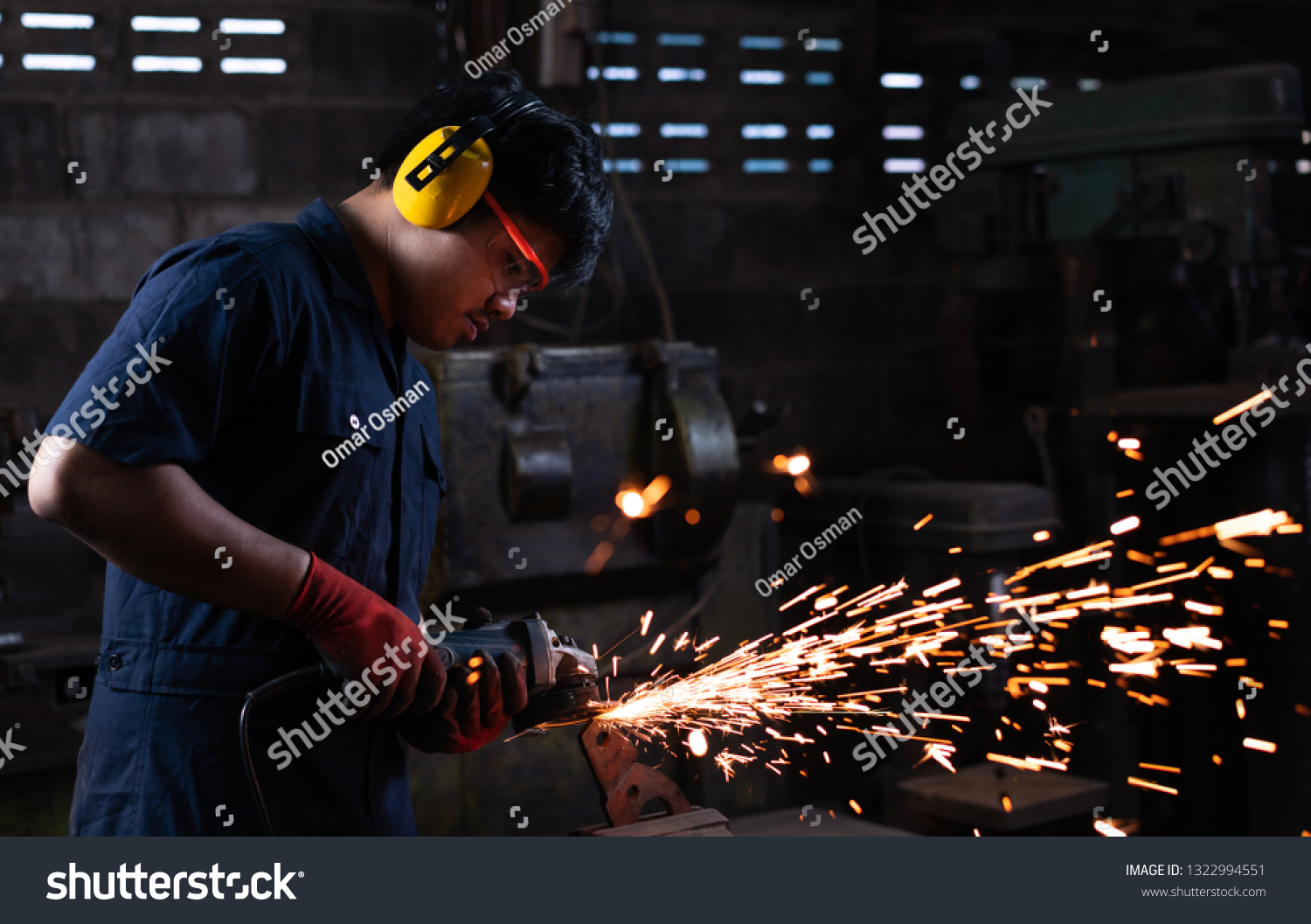 Asian mechanical engineer worker wearing safety equipment and operating angle grinder - Metal factory employee working with power tools in dark industrial workshop with flash sparks - Industry concept #1322994551