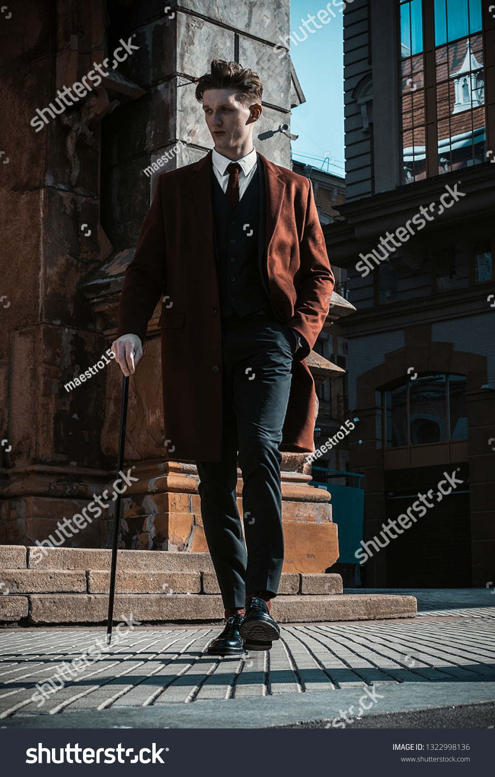 Fashion portrait of young man on red coat, white shirt, black suit and cane walking on streets of city background. Model shooting #1322998136