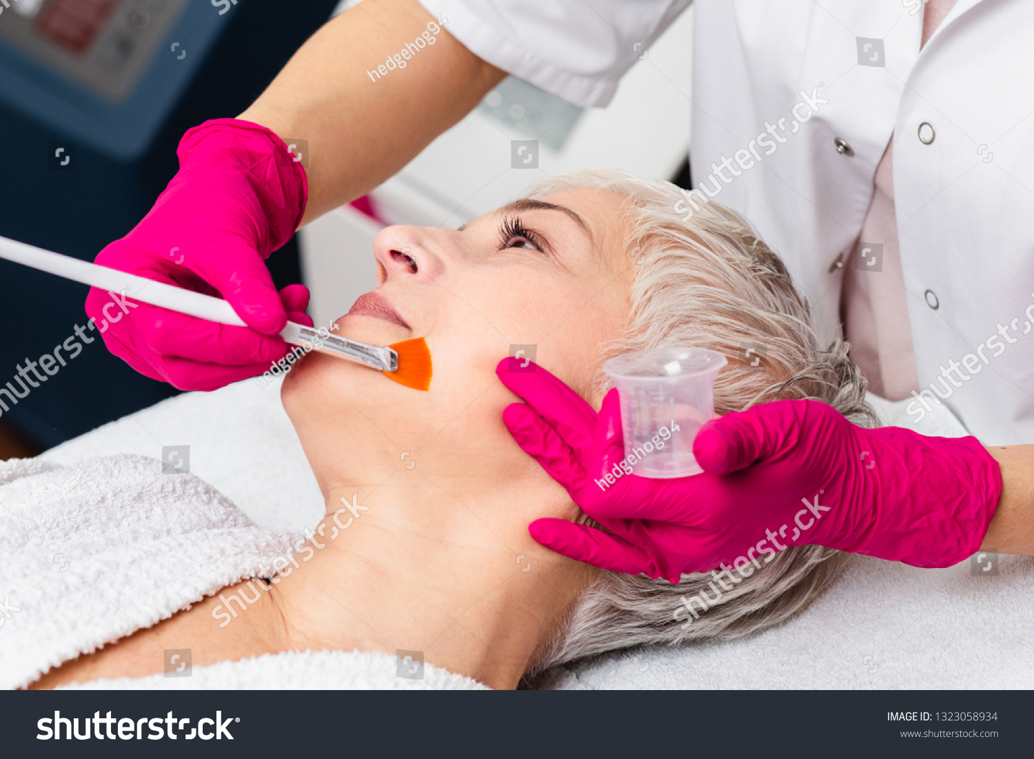Beautiful senior woman having chemical peeling beauty treatment. The expert beautician is applying chemical solution onto woman's face. #1323058934