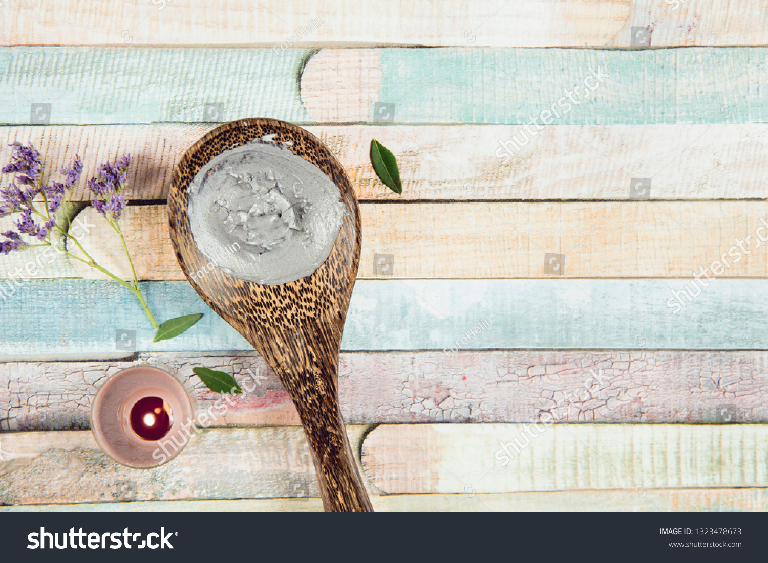 Natural gray mud mask cream on wooden spoon with candle burning and flower branch as decoration on colorful wooden background. Flat lay view, copy space, copy space. #1323478673