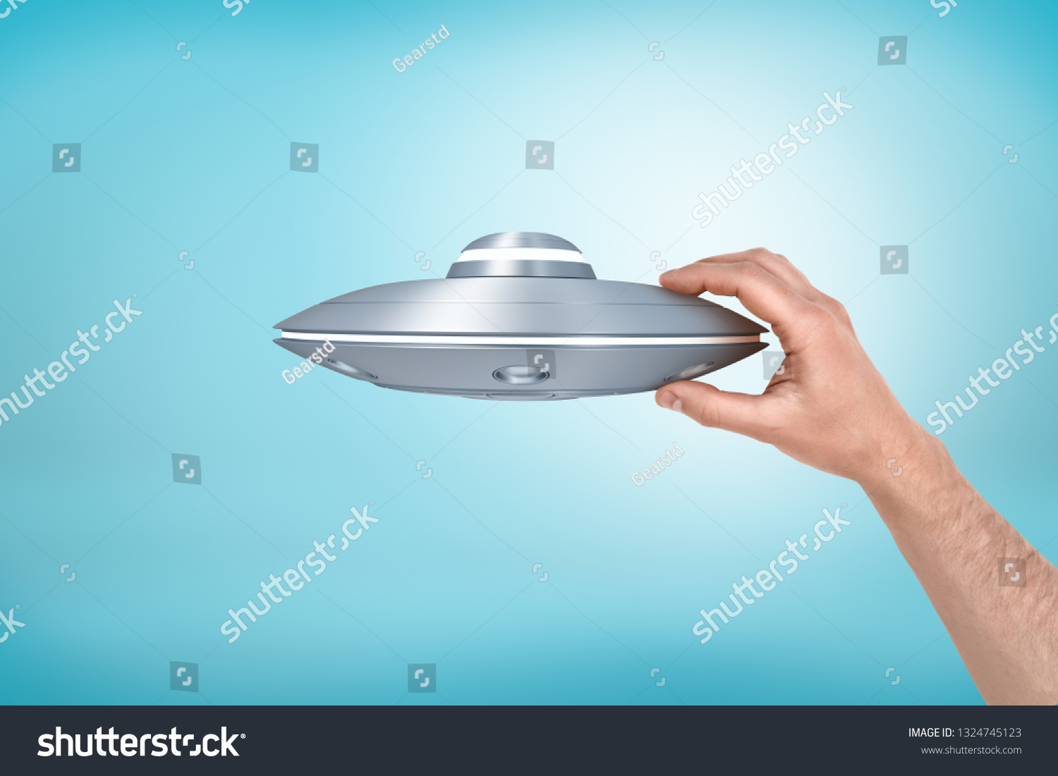 Close-up of man's hand holding small light-gray UFO on light-blue background. Ufology club. Close encounter. I want to believe. #1324745123