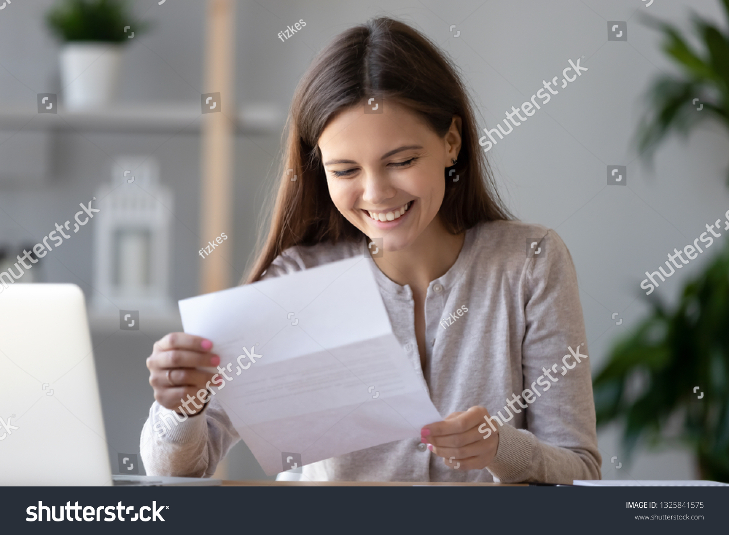 Smiling happy woman student or worker reading good news in paper letter, sitting at desk with laptop, receive great job offer, success exam results, holding document in hands, great deal #1325841575