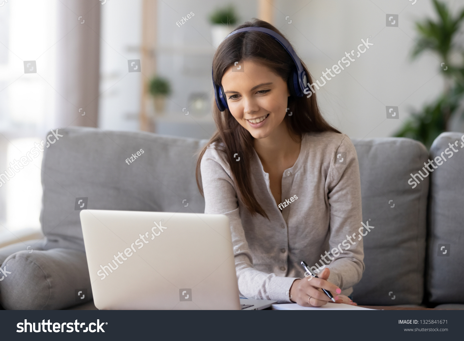 Smiling woman in headphones using laptop, writing notes, sitting on sofa at home, happy girl looking at computer screen, watching webinar, learning language online, distance education concept #1325841671