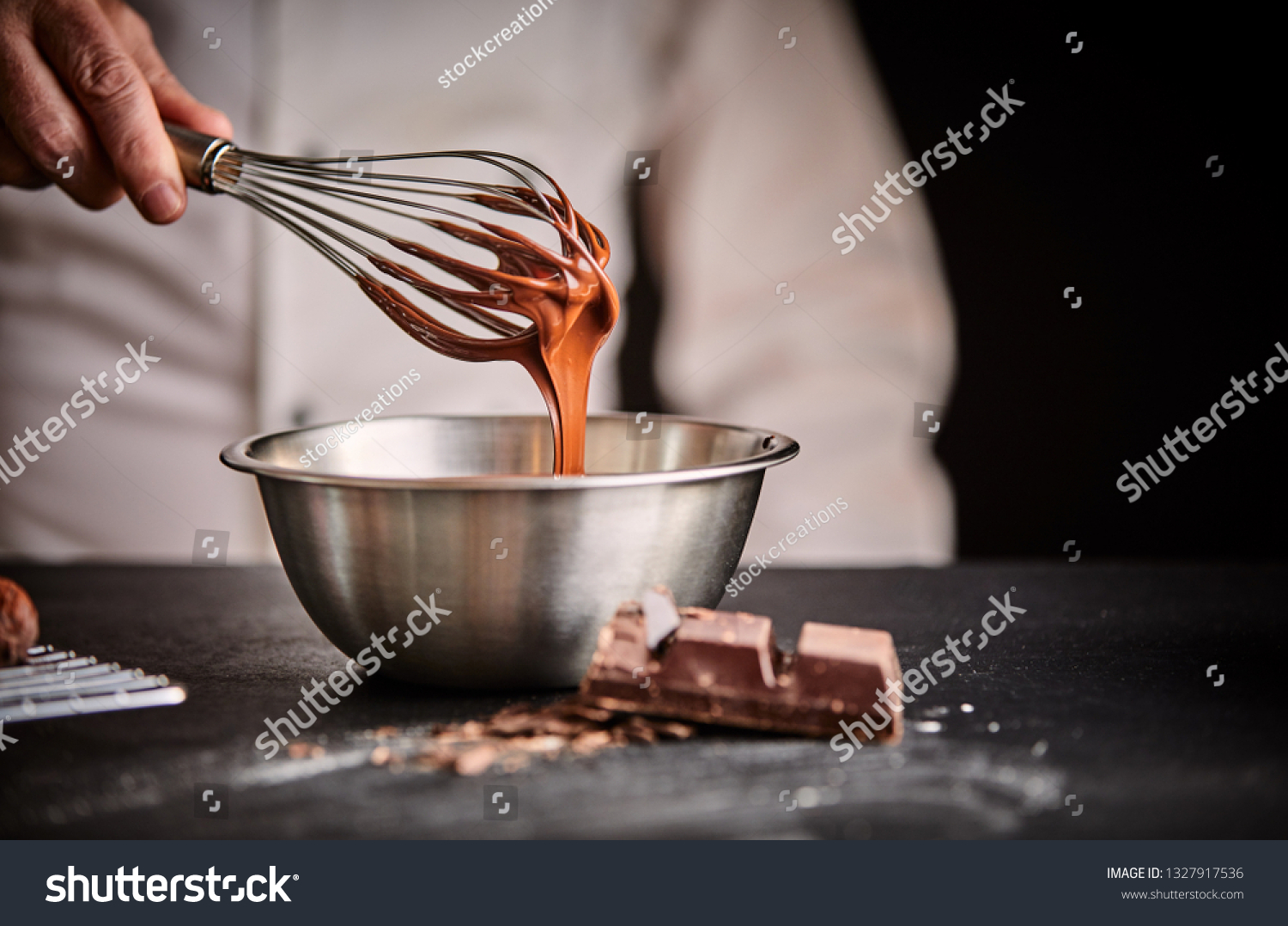 Chef whisking melted chocolate in a stainless steel mixing bowl using an old vintage wire whisk in a close up on his hand #1327917536