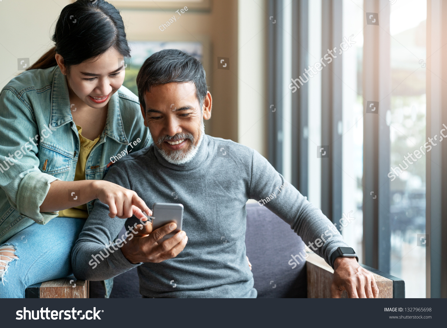 Attractive mature asian man with white stylish short beard looking at smartphone computer with teenage eye glasses hipster woman in cafe. Teaching internet online or wifi technology concept #1327965698