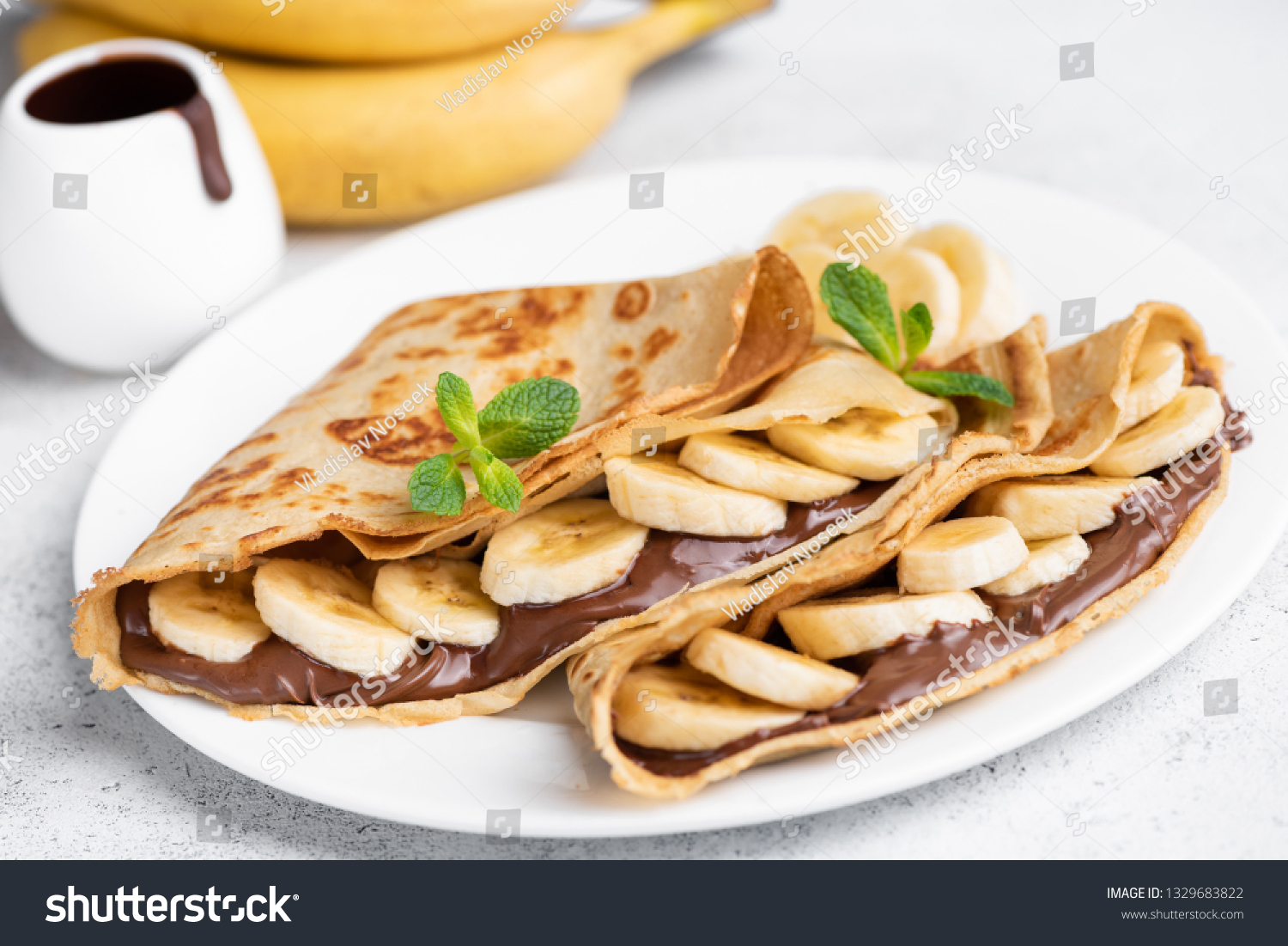 Crepes stuffed with chocolate spread and banana on white plate. Thin pancakes, blini. Sweet dessert. #1329683822