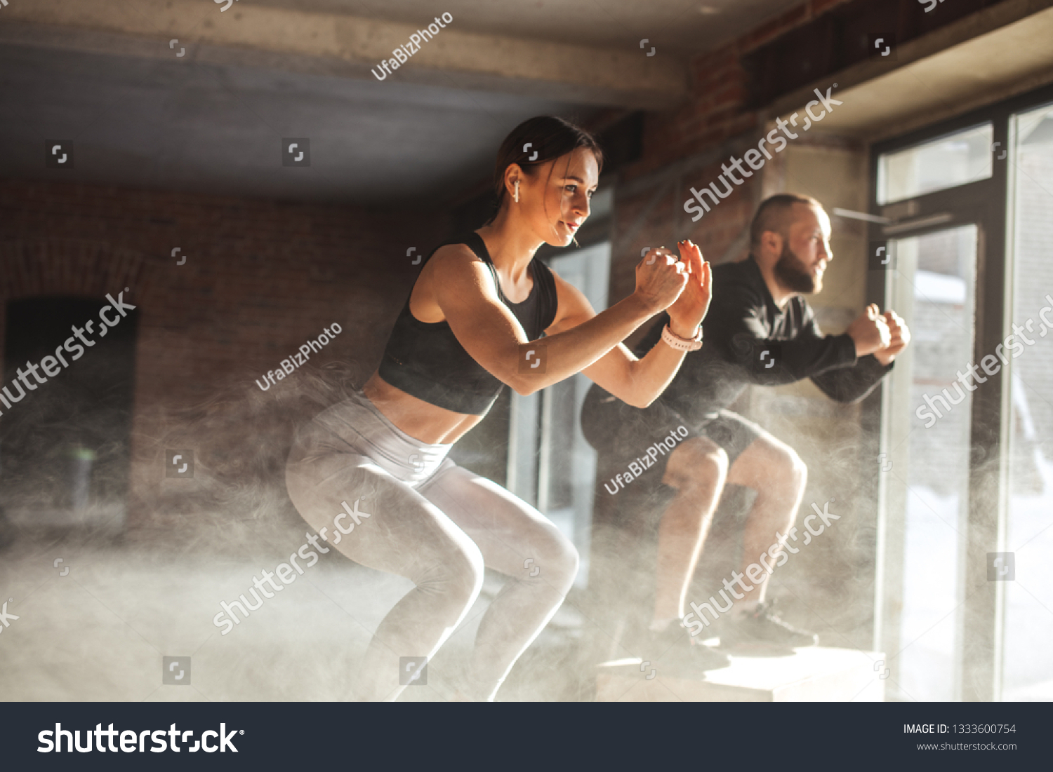 Fit caucasian couple doing squats on boxes in a crossfit style gym, well lit by bright sunlight. #1333600754