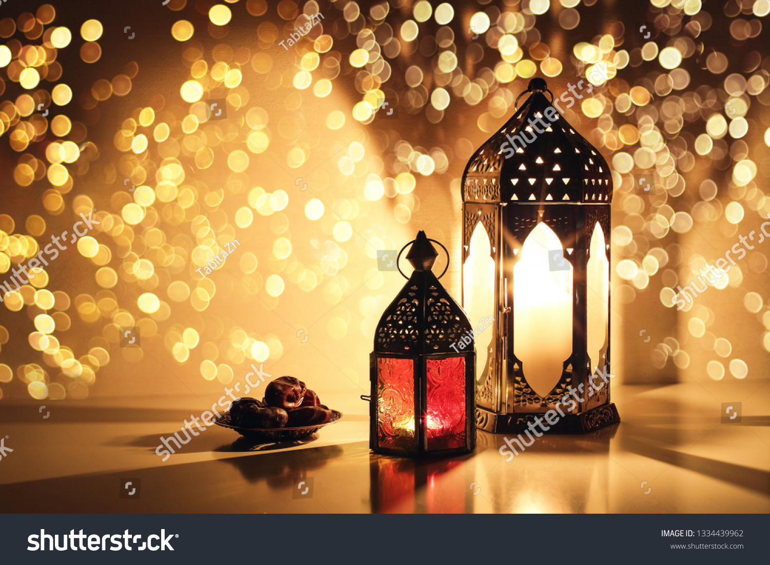 Ornamental Arabic lanterns with burning candles. Glittering golden bokeh lights. Plate with date fruit on the table. Greeting card for Muslim holiday Ramadan Kareem. Iftar dinner background. #1334439962