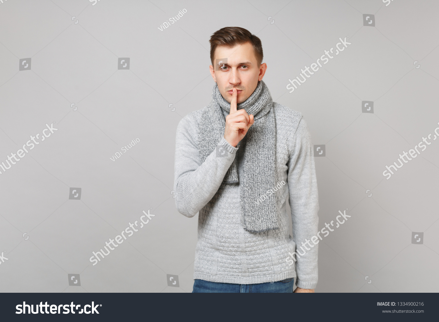 Young man in gray sweater, scarf saying hush be quiet with finger on lips shhh gesture isolated on grey background. Healthy lifestyle, people sincere emotions, cold season concept. Mock up copy space #1334900216
