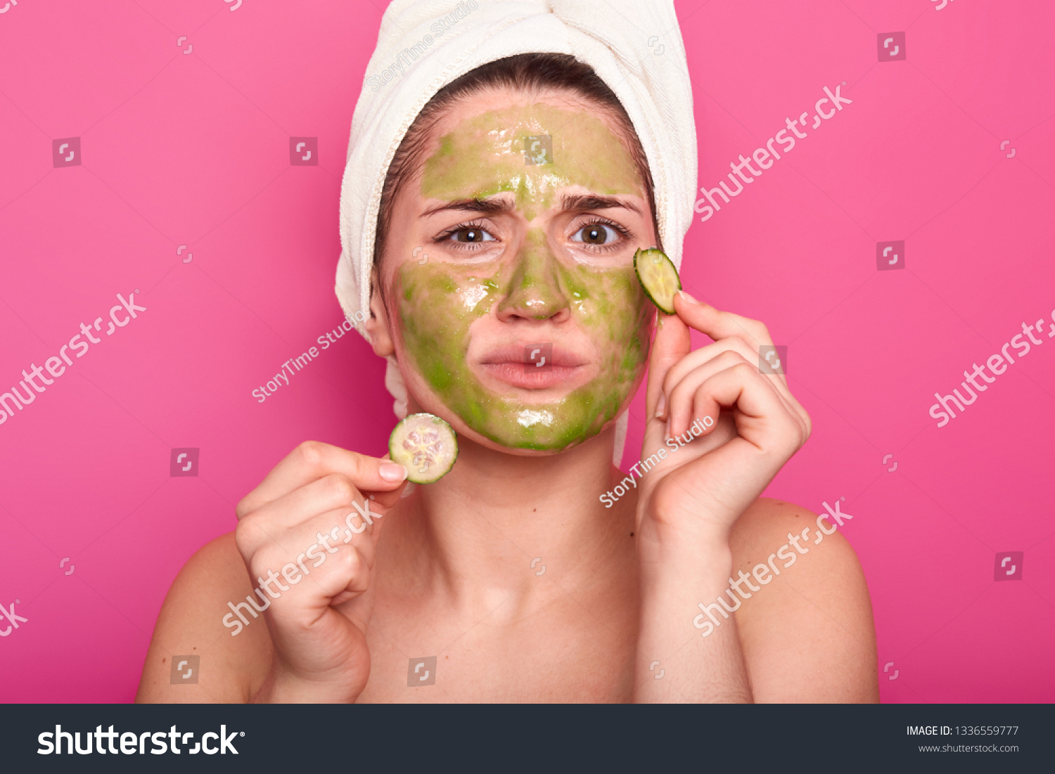 Dissatisfied young cute woman doesnt like smell from beauty mask, discontent with effect, holds two slices of cucmber, removes wrinkles on face, wears white towel, isolated on pink studio wall #1336559777