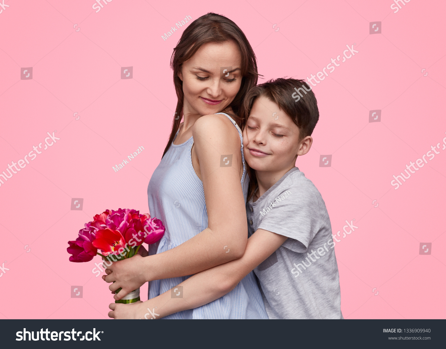 Side view of boy embracing mother from back while congratulating with 8 March and giving bouquet of tulips on pink backdrop  #1336909940
