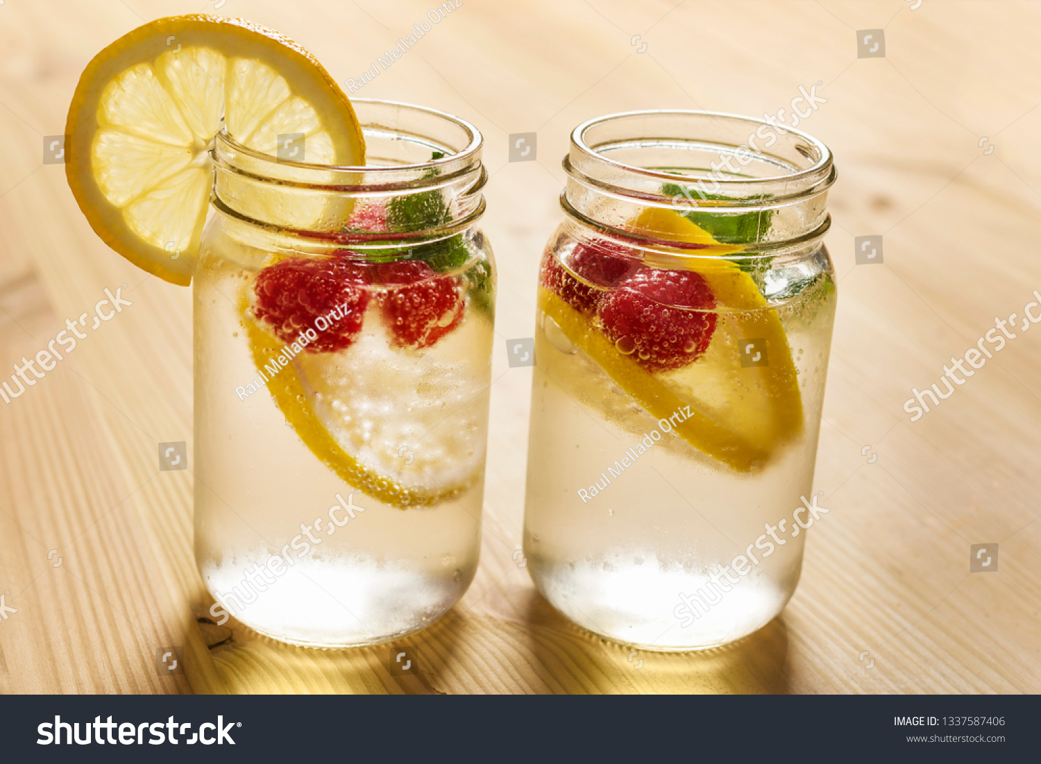 two glass jars with cold water, slices of lemon, mint and red berries, illuminated by sunlight on a wooden table with some pieces of citrus, summer refreshments background, copy space #1337587406