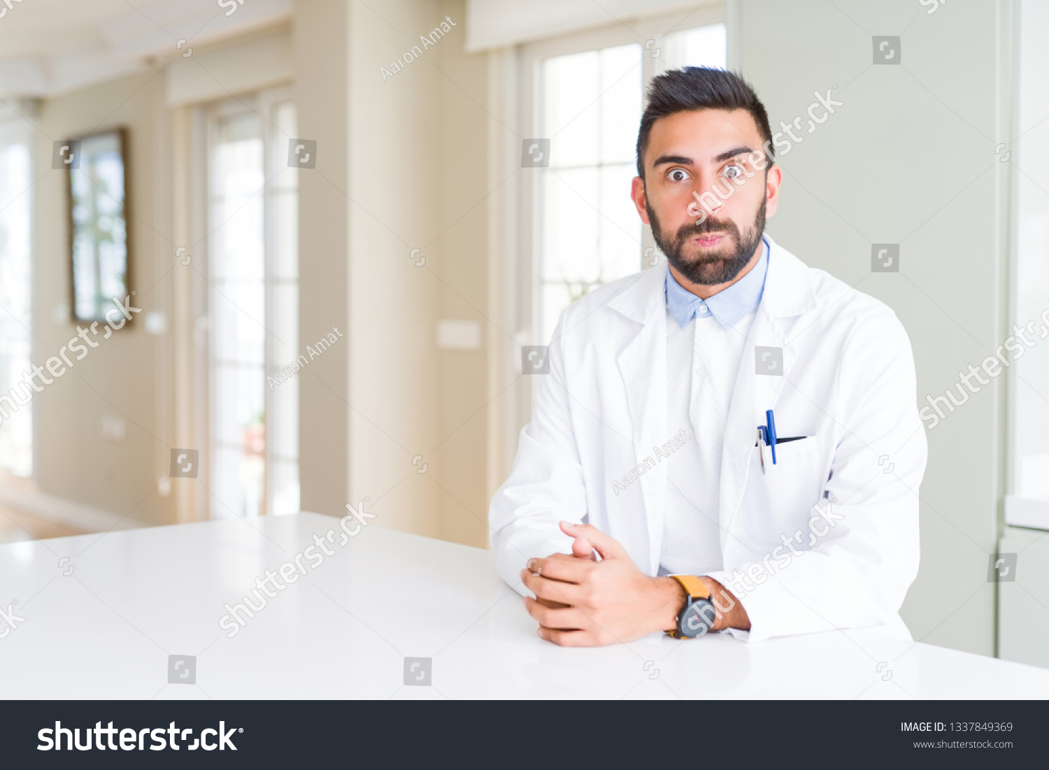 Handsome hispanic doctor or therapist man wearing medical coat at the clinic puffing cheeks with funny face. Mouth inflated with air, crazy expression. #1337849369