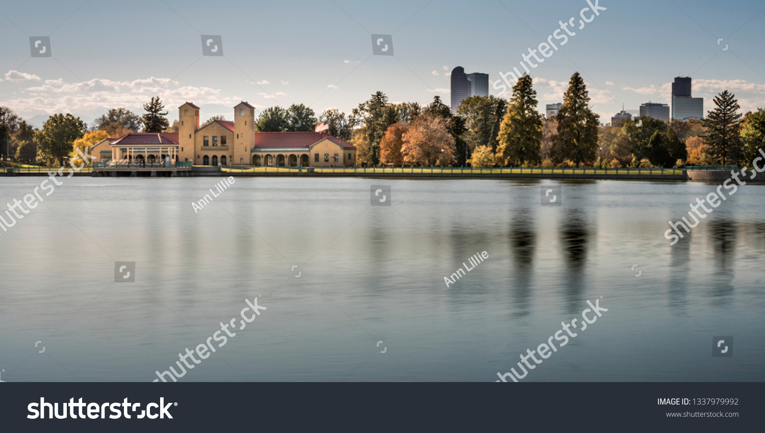 A beautiful panorama of City Park in Denver, Colorado in late summer or autumn. The brick boathouse and a row of trees changing color are reflected in the smooth, blue, peaceful water of the pond. #1337979992
