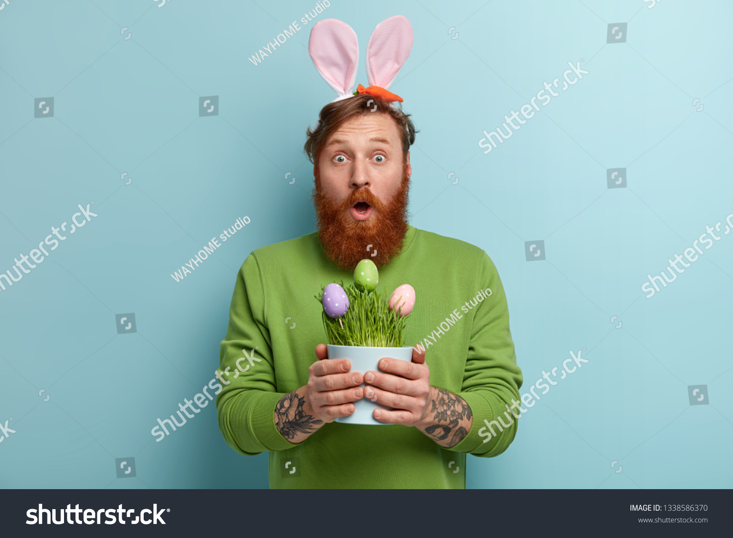 Stupefied bearded adult man wears bunny long ears on head, carries Easter symbols, opens mouth from wonder, holds pot with colored eggs, wears green sweater, poses indoor. Spring time concept #1338586370