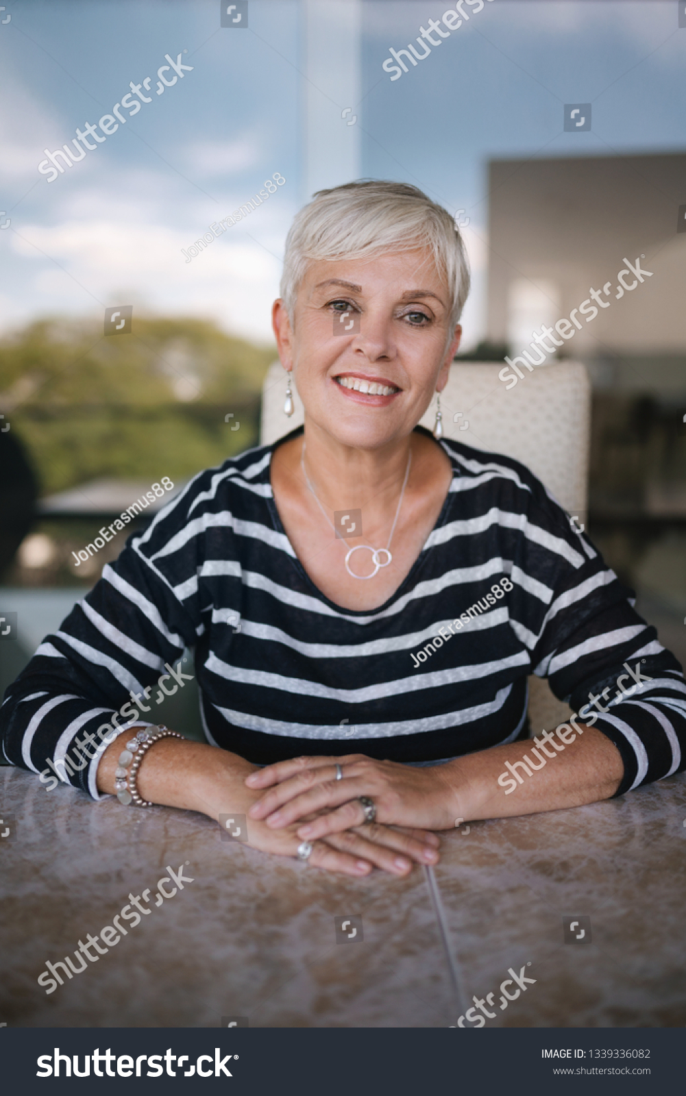 Beautiful elderly woman smiling looking into the camera. Portrait of a mature woman with hands crossed on the table, outside on the balcony. Beautiful elderly woman smiling looking into the camera #1339336082