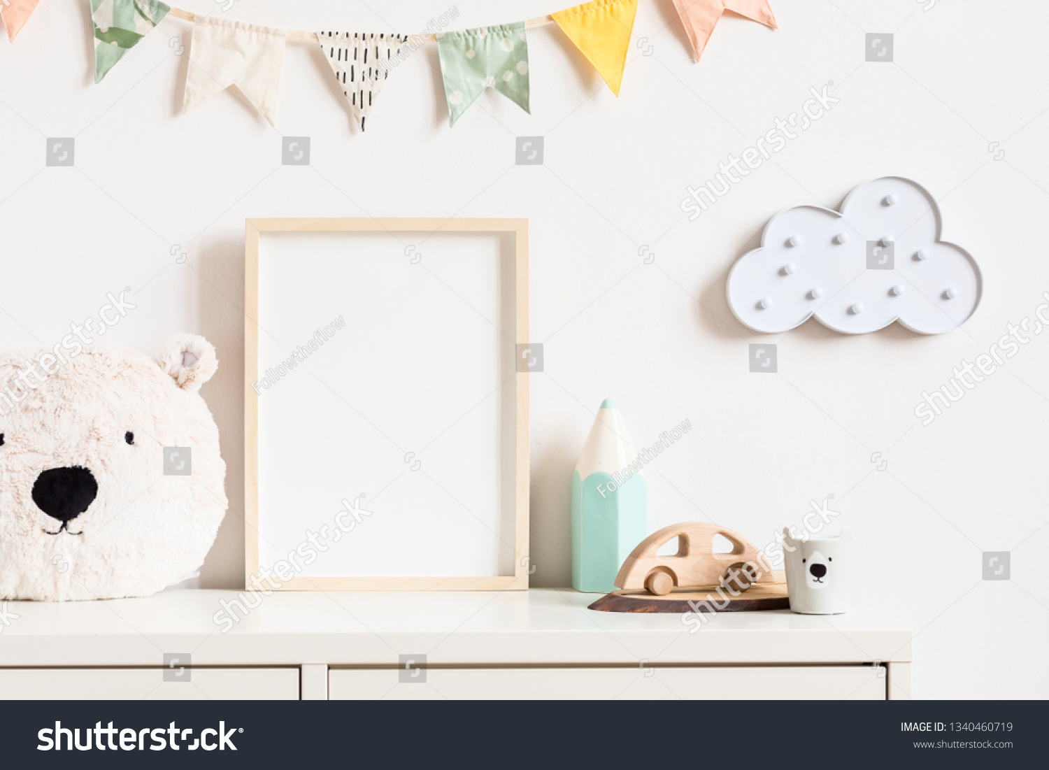 Stylish and modern scandinavian newborn baby interior with mock up photo or poster frame on the white shelf. Toys, teddy bear, wooden car and hanging cotton colorful flags and star. Template. Blank. #1340460719