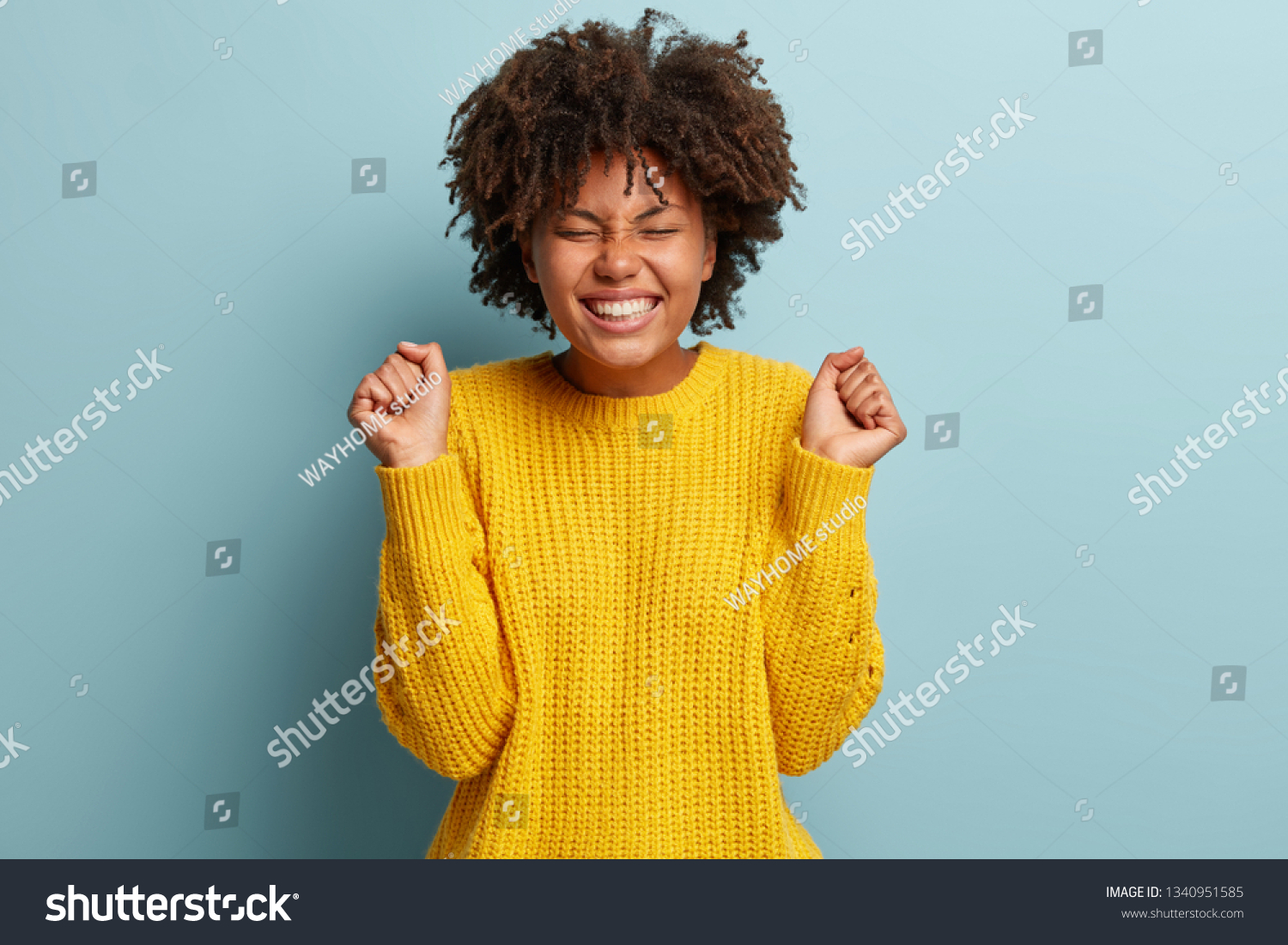 Overemotive triumphing woman squints face from excitement, clenches fists in victory, hears amazing good news, shows broad smile, wears oversized yellow jumper, gestures joyfully. Success concept #1340951585