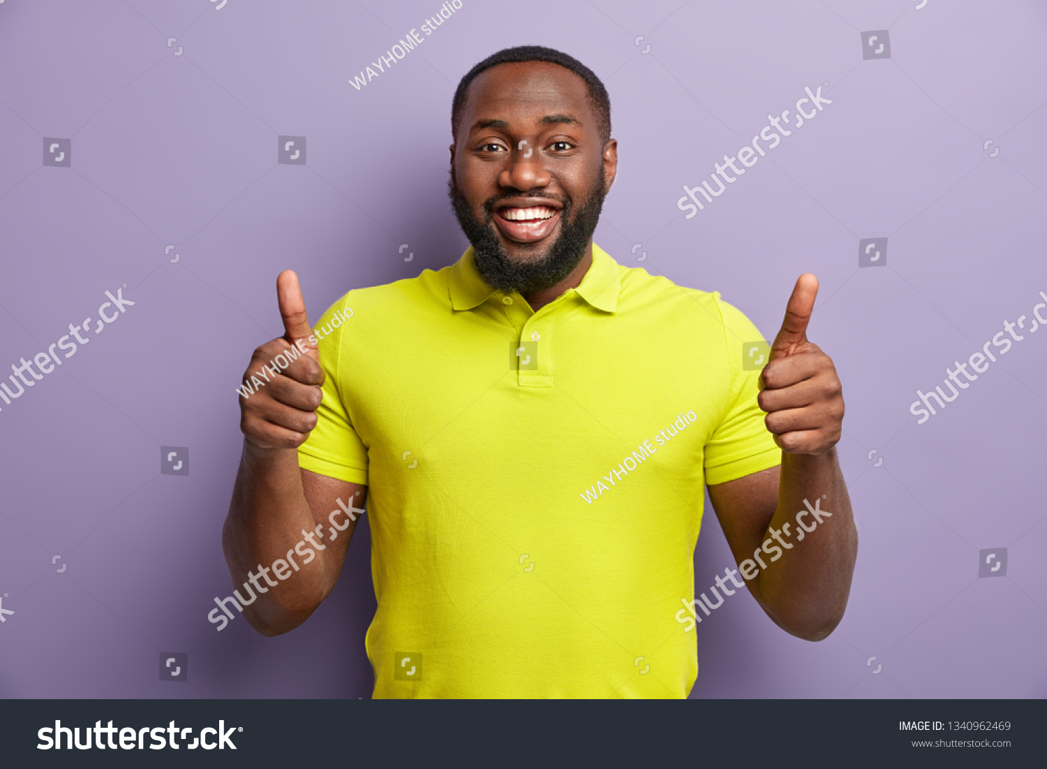 Satisfied black man gestures with both hands, shows approval gesture, keeps two thumbs raised, smiles broadly, wears yellow casual t shirt, stands against purple background. Thats nice, I like it #1340962469