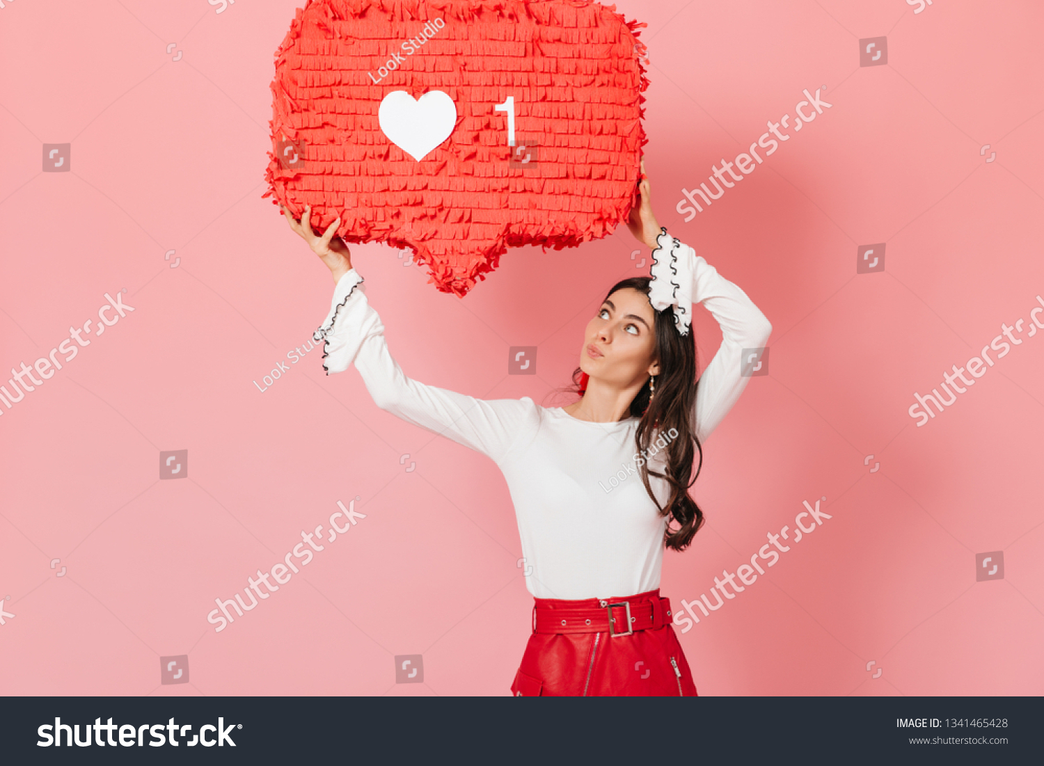 Portrait of girl with interest looking at huge ""like"" sign from Instagram. Brunette in red skirt posing on pink background #1341465428